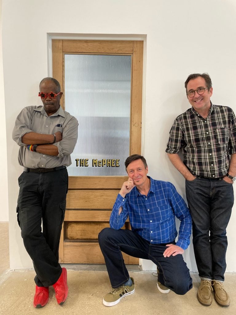 Three men stand in front of a door with the name "The McPhee" printed on the glass