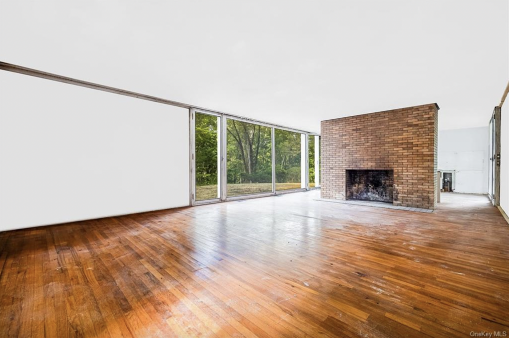 A photo of the sparse interior of Philip Johnson's Booth House, with wooden floors, a white ceiling, a brick fireplace, and floor to ceiling glass windows.