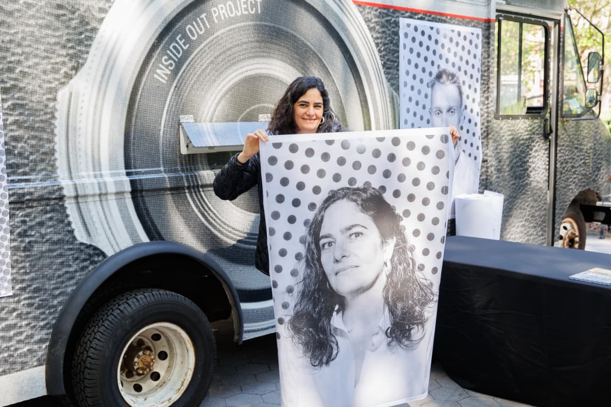 A woman with her portrait, taken by JR's "Inside Out Project" photo booth truck for "Portraits on Climate and Health: Dreams We Carry" at the American Museum of Natural History in New York. A woman with long dark hair holds up a black and white portrait of herself against a polka dot background that is nearly as tall as she is, standing in front of a truck that has a massive camera lens printed on its side.