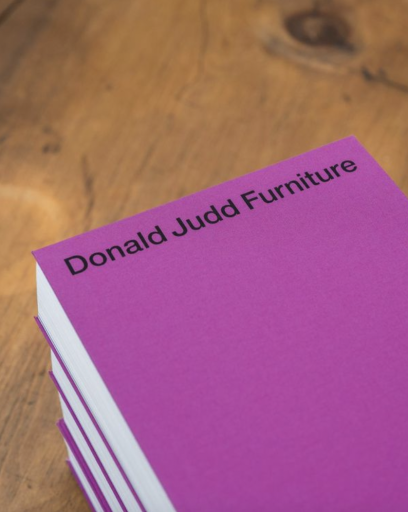 A photograph of a plain purple book labeled "Donald Judd Furniture" in black text at the top, on a warm brown wooden table