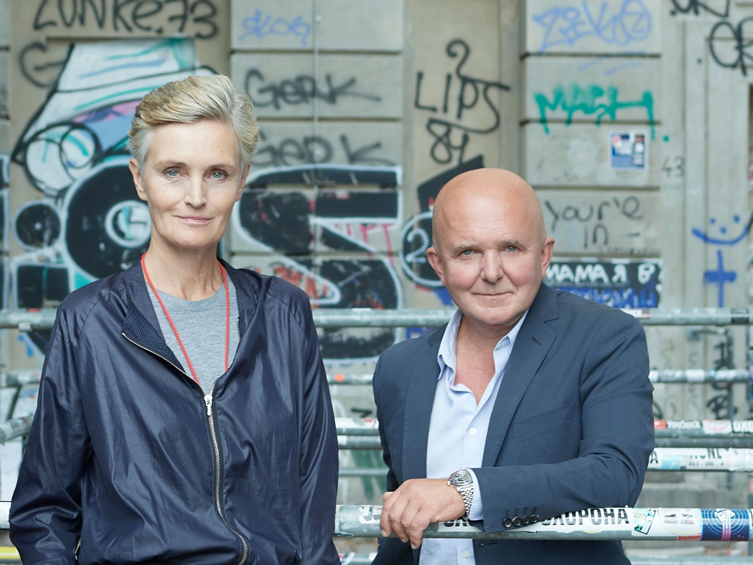 On the left Karen Boros in a blue windbreaker and gray crewneck, on the right Christia Boros wearing a navy suit jacken at light blue button down and silver watch, both in front of an industrial style wall with graffiti. Both are the recipients of the Art Cologne Prize.