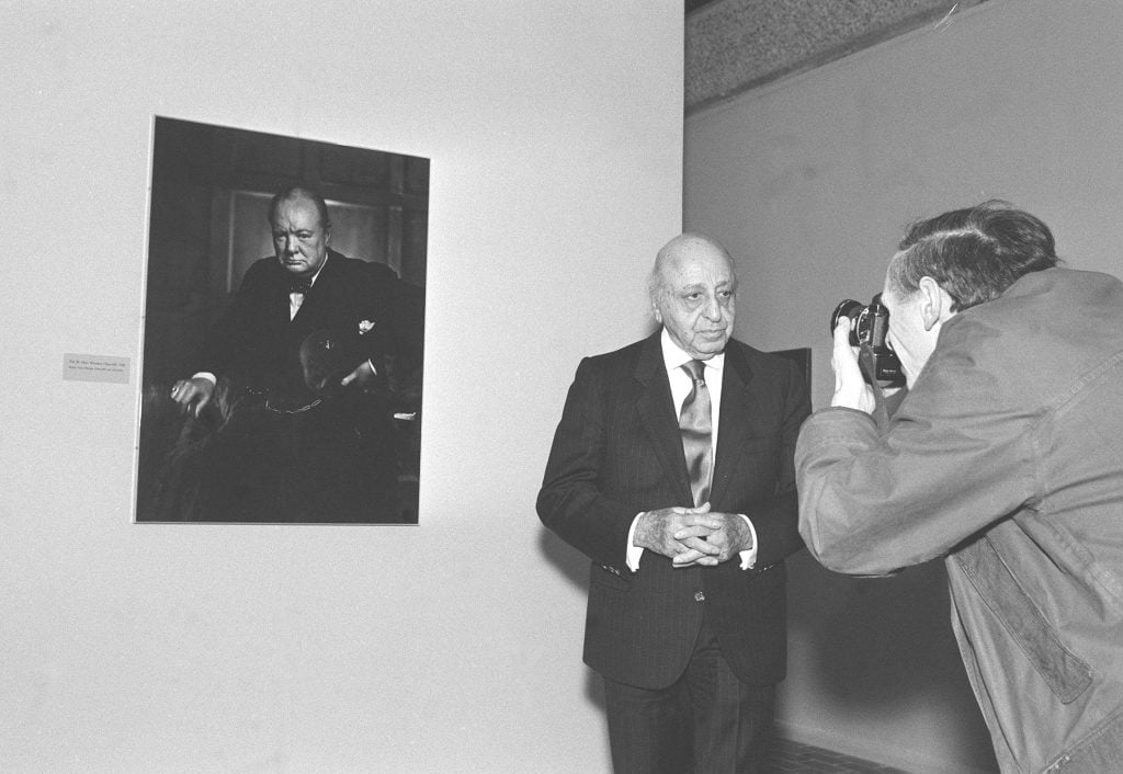 A black and white photograph of Yousuf Karsh having his photo taken in front of his famous portrait of Winston Churchill, hanging on a white wall behind him.