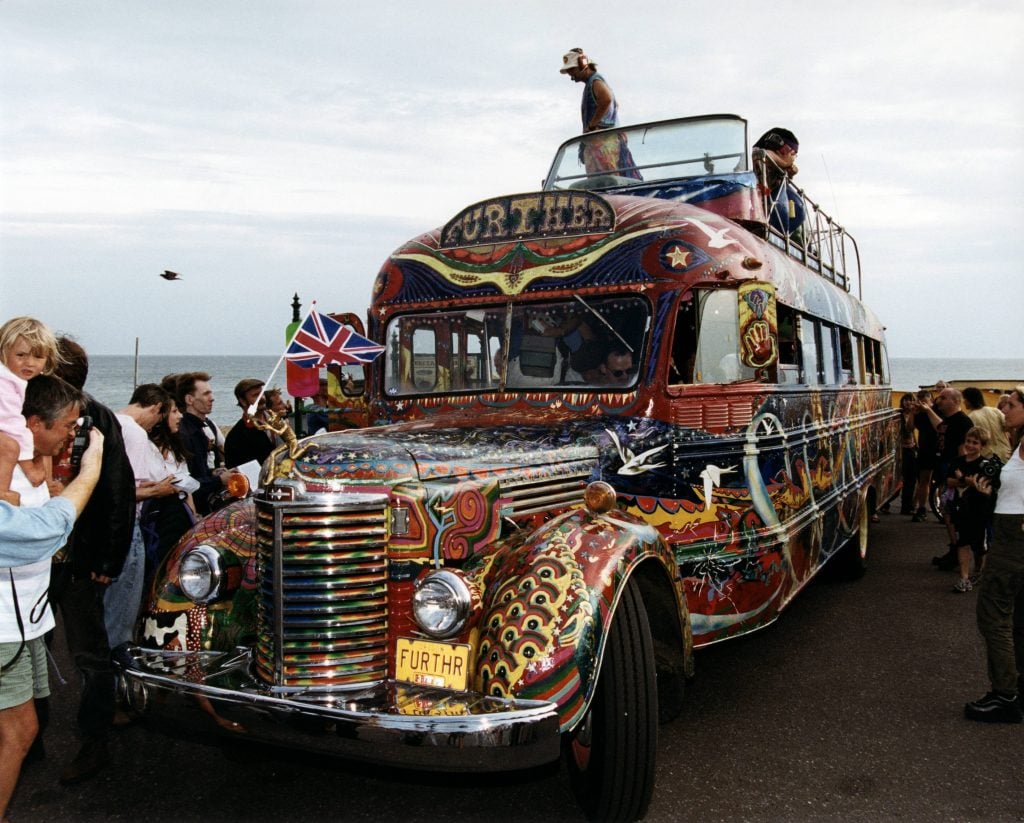 Ken Kesey and the Merry Band of Pranksters' bus, Further, a school bus painted in bright colors, by the waterside in Brighton greeted by a crowd of people. Two people are on the roof of the bus, which has a Union Jack flag flying on the front.