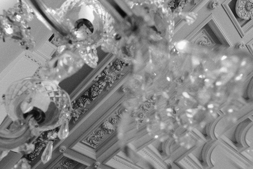 A black and white detail photo of a crystal chandelier and intricately decorated plaster ceiling.