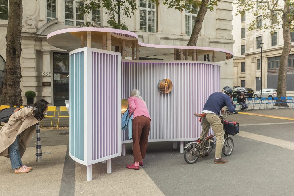 A photo of a curved, midcentury model-style installation in lavender on a city street, with passersby peering intently into the circular peepholes that dot it.