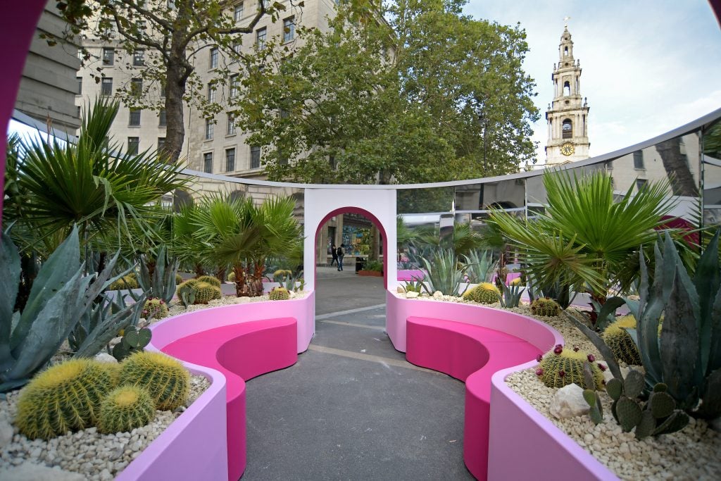 A photo of an empty, open air courtyard pavillion in a city square which features curved, mirroed walls and pink shelves lined with cacti.