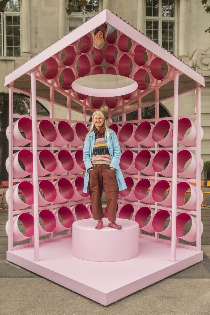 A photo of a woman standing under the mirrored roof of a pink geometric art installation, with a light pink stool in its center.