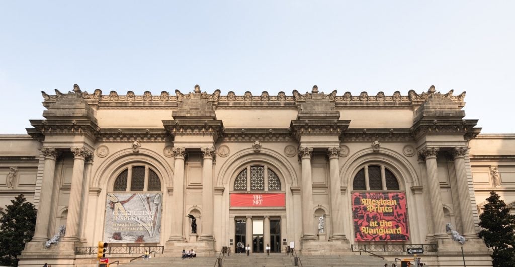 A photograph of the Met's main entrance beneath light blue skies, holding four sculptures by Lee Bull in its exterior facade niches.