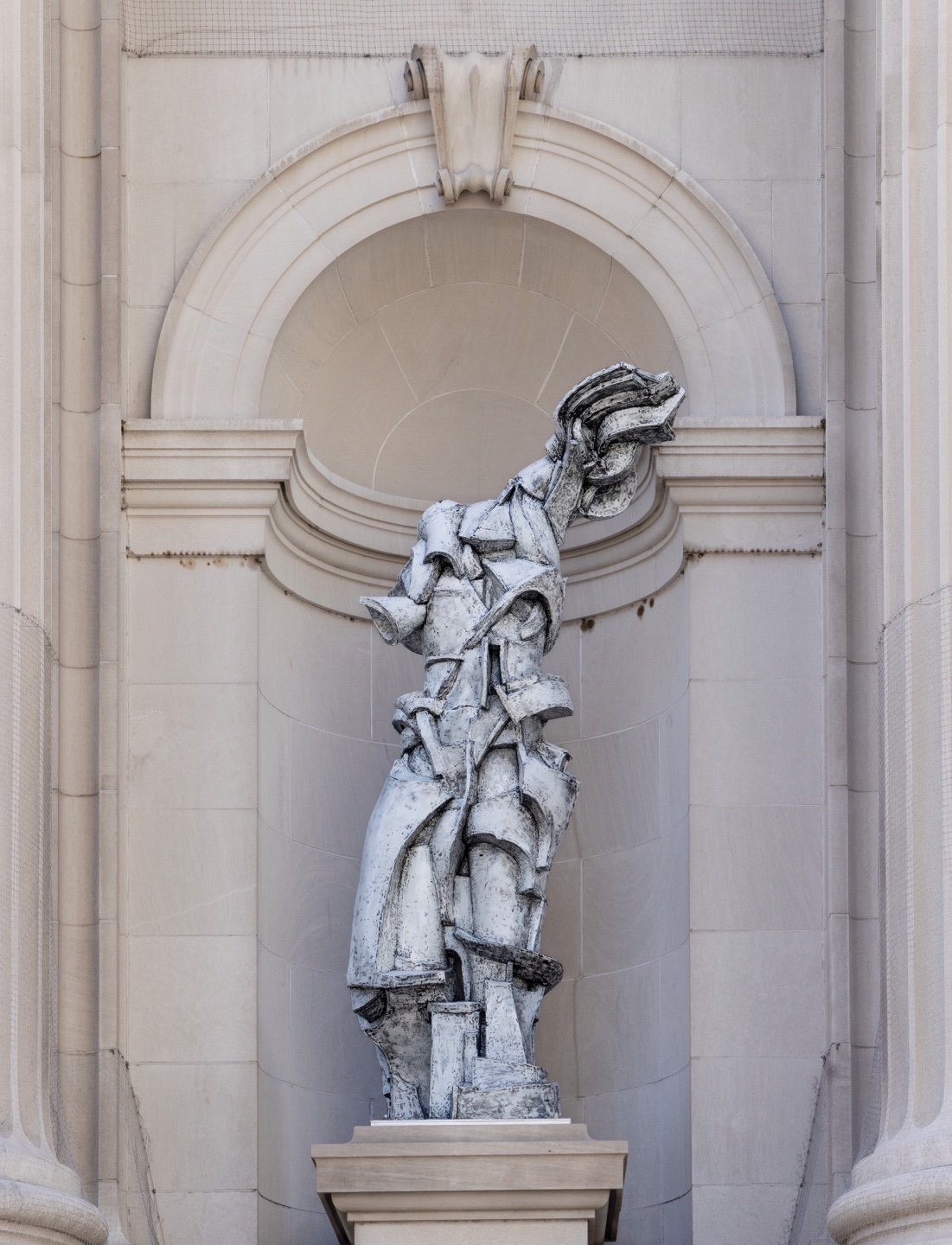 A photograph of a white abstract sculpture by Lee Bul loosely resembling a woman in a niche on the facade of the Met.