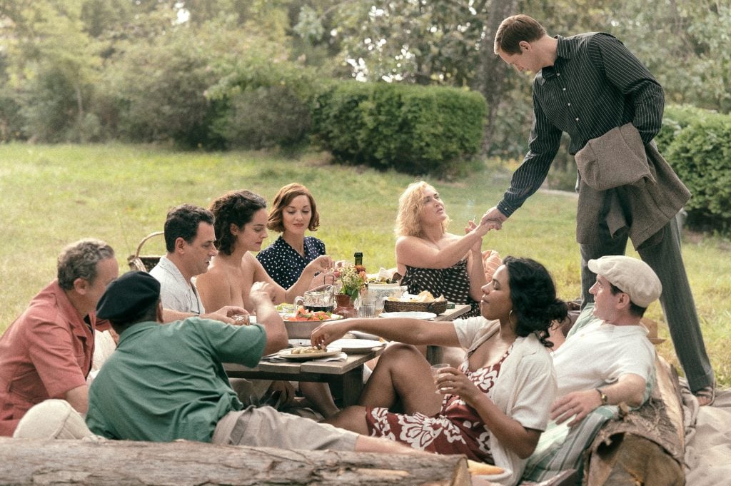 A group of men and women are dining outdoors. A woman extends her hand to shake a standing man.