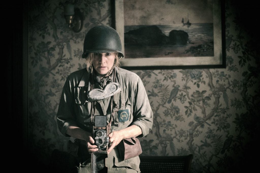 A woman in combat gear holding a Rolleiflex camera in a dusty wallpapered room