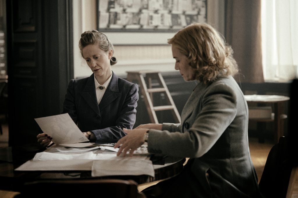 Two suited women sitting at a table covered in books and papers