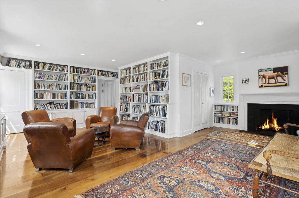 A photograph of a white living room lined with books, featuring leather chairs, a pattered rug, and a lit fireplace.