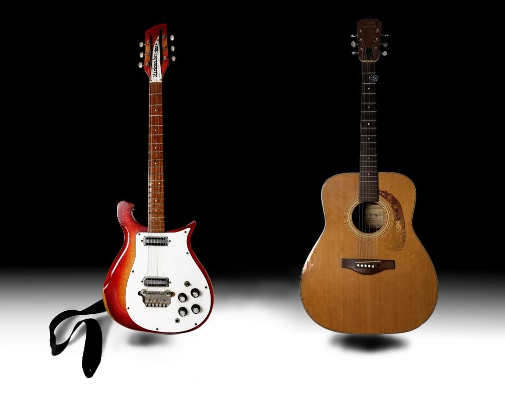 A photograph of red and white electric guitar next to a brown acoustic guitar, both on stands atop a white floor on a black background.