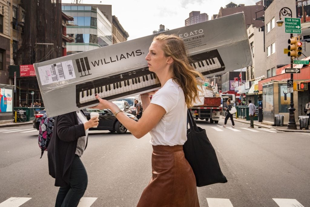 A woman walking across a crosswalk carrying over her shoulder an electric keyboard, wearing a white tee and brown skirt.