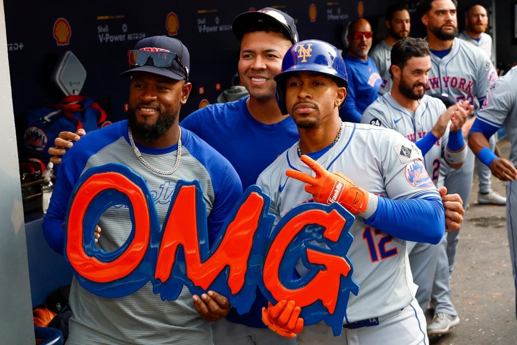 Francisco Lindor (right), shortstop of the New York Mets, celebrates with pitchers Luis Severino (left) and Jose Quintana (center) with an OMG sign by artist Jerome McCroy after hitting a solo home run in the ninth inning during a game against the Toronto Blue Jays on September 11, 2024, in Toronto, Canada. The players are in the dugout, and the sign is large in bright blue and orange.