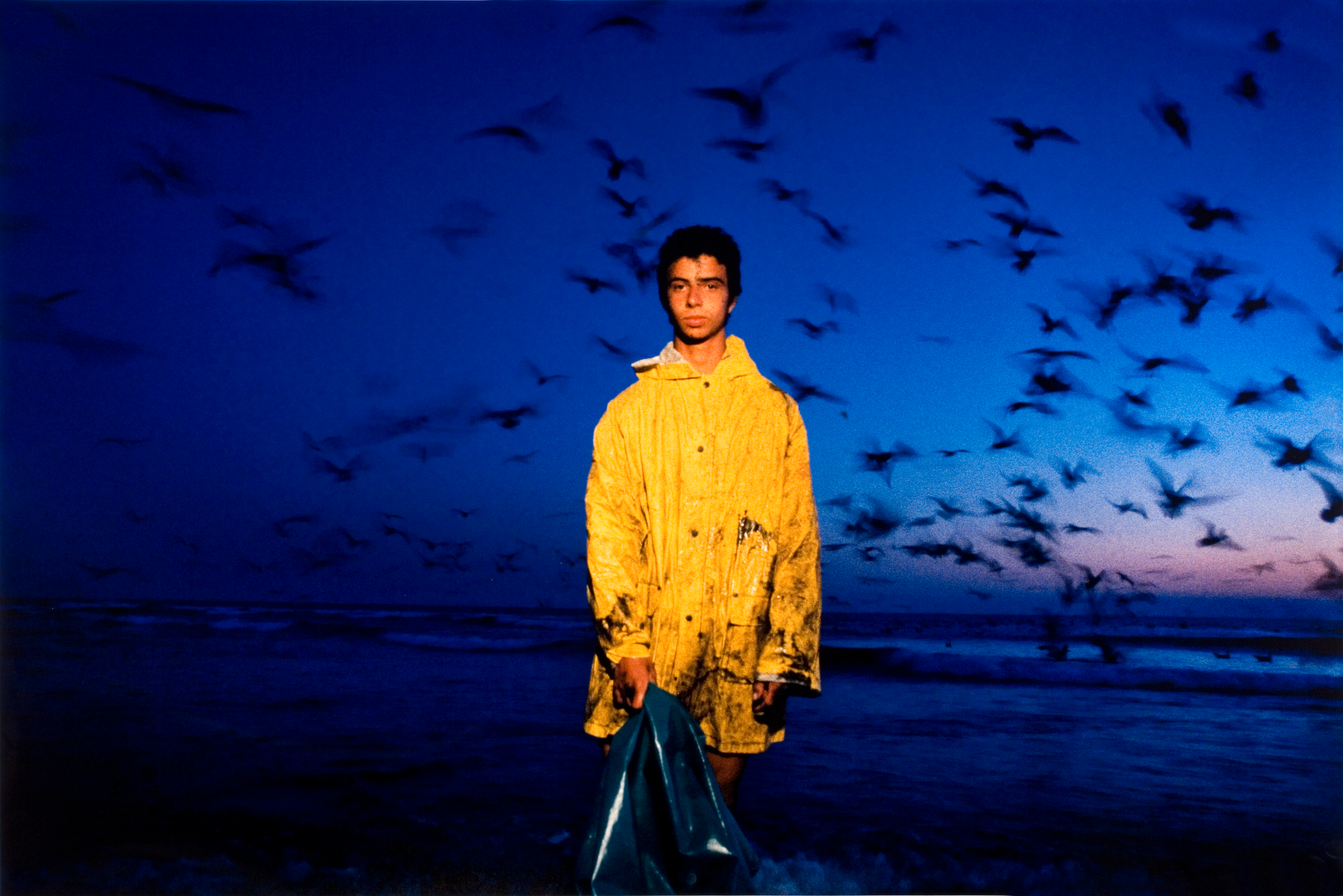 A young man in a yellow raincoat stands before a vivid blue sky