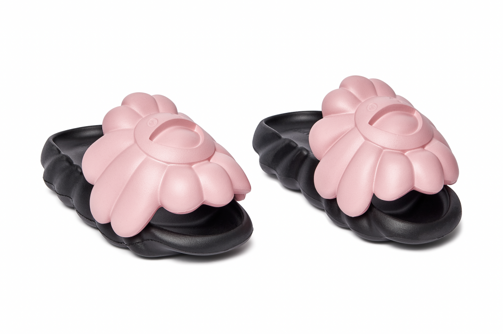 A photo of a pair of black slide-on flip flops, with soft pink top straps in the shapes of Takashi Murakami's smiling flowers.