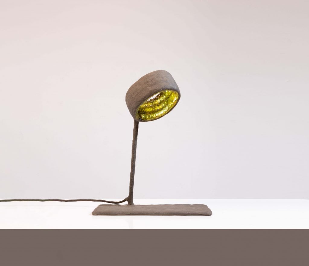 A photo of a simple lamp made of sand with green crystals visible from within its shade sitting atop a white table, before a grey backgroud.