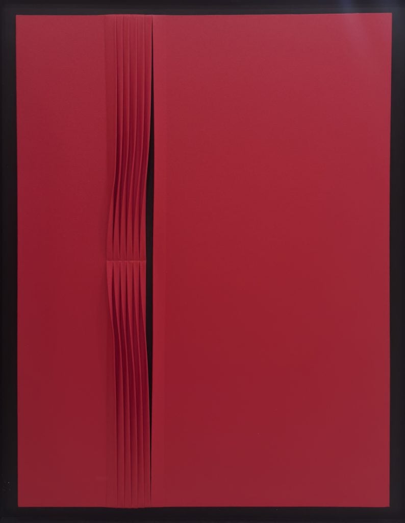 A red monochrome painting with a vertical slit and a series of slim pleats to the left. Featured in the the group show forms in flux.