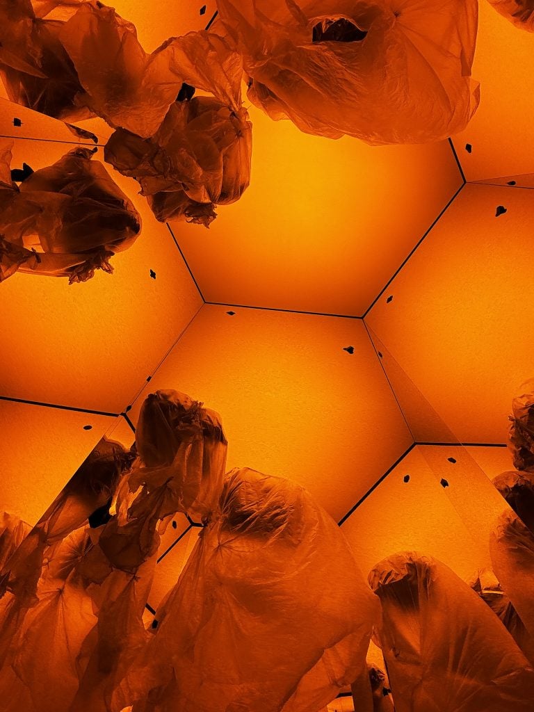 An orange-tinged installation with a roof made up of hexagonal tiles and floating plastic bags