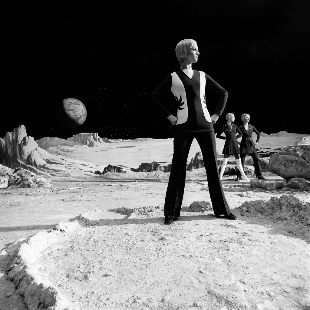 A black and white image of a woman in 1970s attire standing on the surface of the moon and the earth in the far black background.