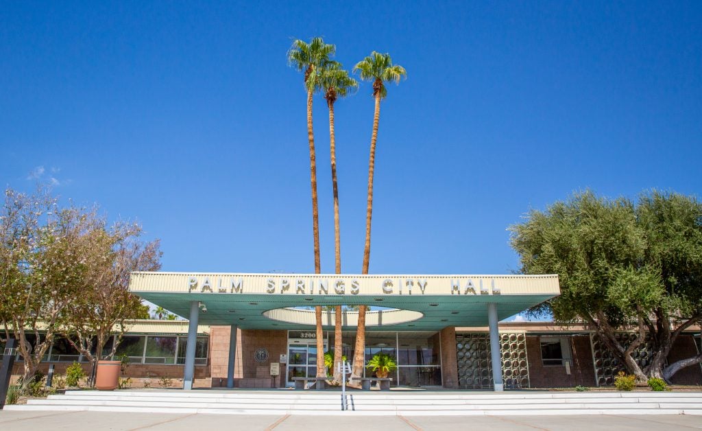 A photo of the mid century modern fa cade of Palm Springs City Hall, with two palm trees front and center, and a brilliant blue sky above