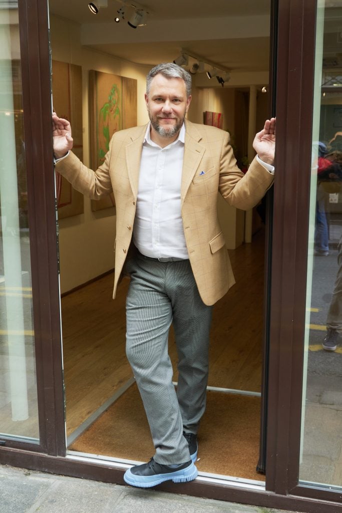 Full-length portrait of Pavel Morozov, wearing blue sneakers, sage pants, a white button down, and light ochre sports coat, standing in the doorway of Galerie De Buci in Paris.