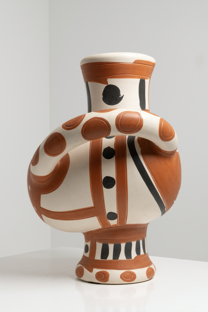 A photo of a white and brown decorated ceramic vase by Pablo Picasso, seen up close in side profile against a grey background.