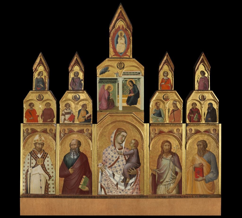 A photo of a large, three-level altarpiece featuring 25 colorfully painted panelsin the Siena Renaissance style, atop a plinth and before a black background.