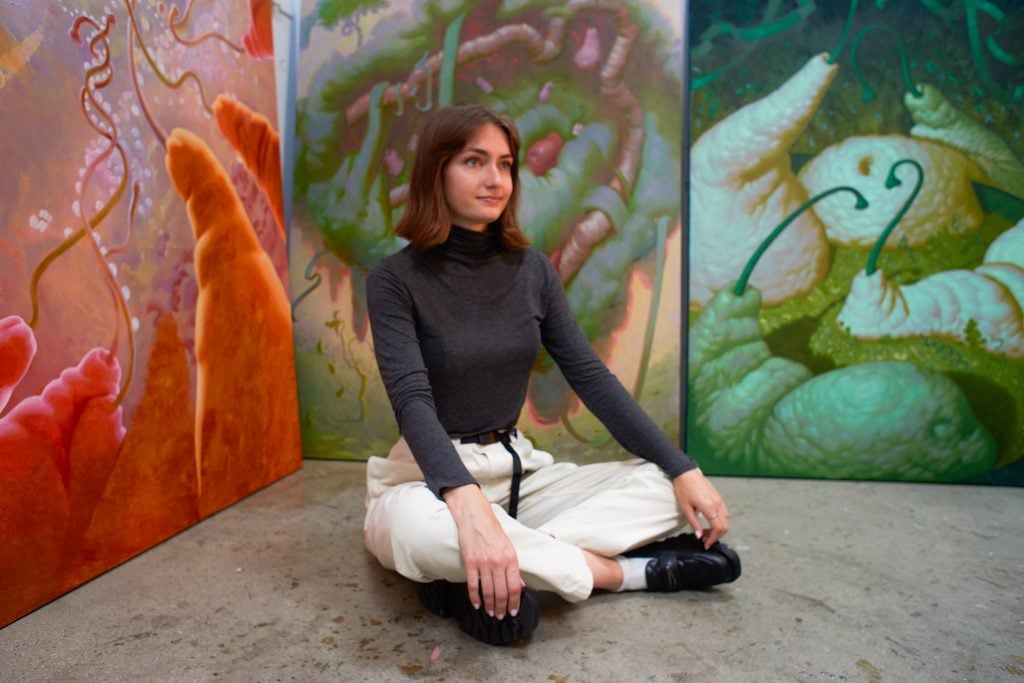 Artist Piper Bangs sitting on the floor of her studio wearing white khakis and turtleneck and black loafers with white socks surrounded by her paintings.