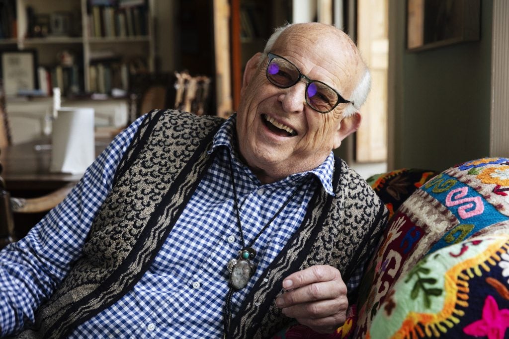 A smiling elderly man in a checked blue shirt reclining on a colorful sofa