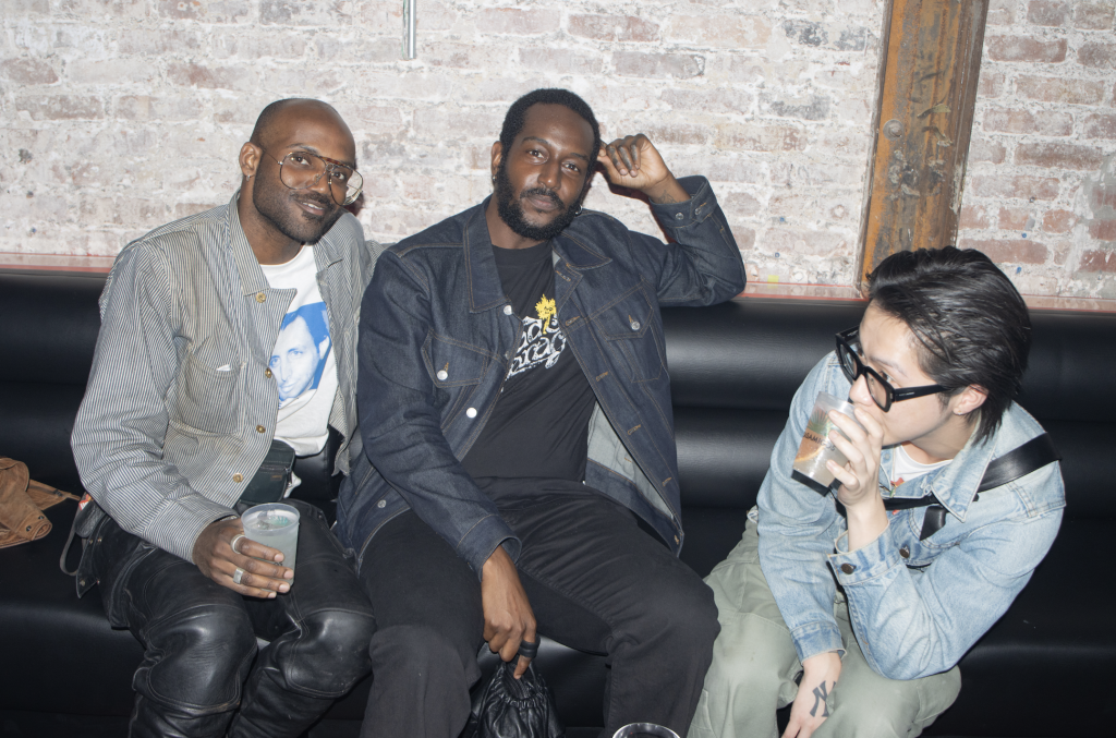 A photo of three partygoers on a black leather couch before a brick wall.