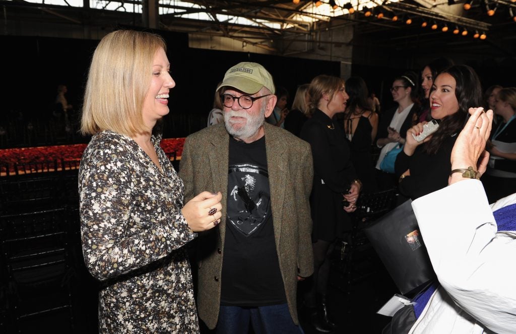 A photograph of an elderly Richard Pettibone in a baseball cap and jacket standing in a crowded room looking at a younger blond woman standing to his left.