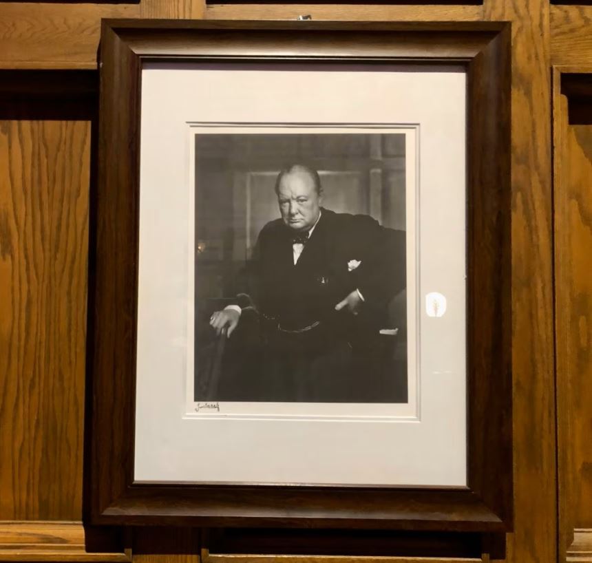 A photograph of a framed and matted black and white portait of Winston Churchill hanging on a wooden wall.