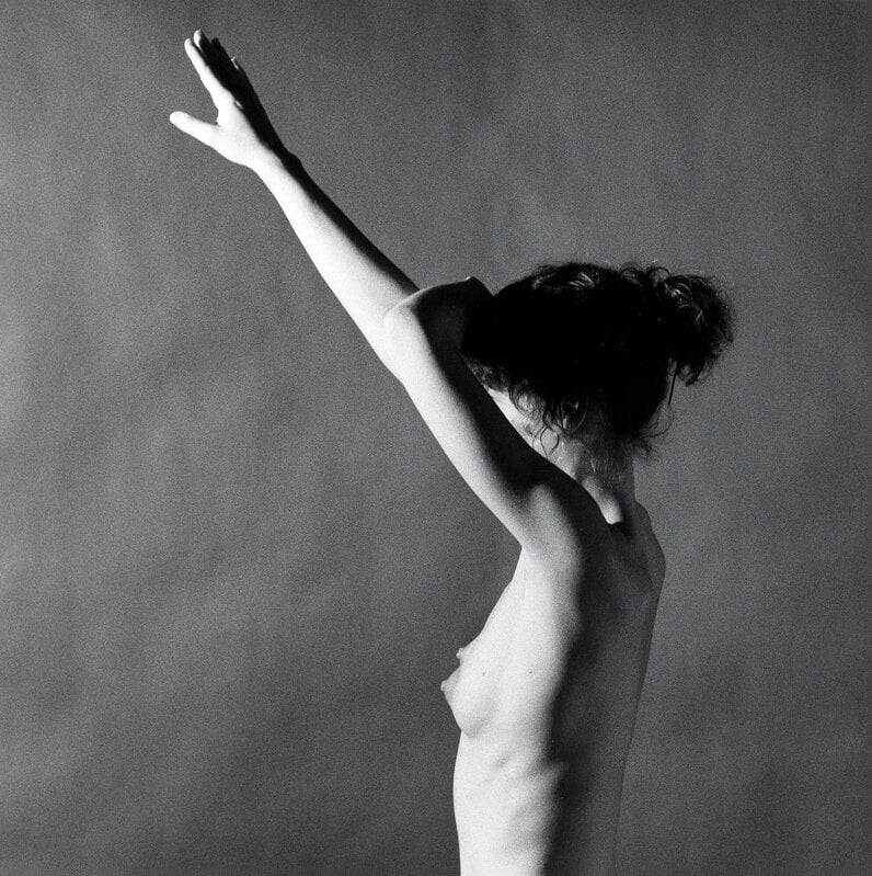 Black and white half-portrait of a nude woman in profile with her arms up obscuring her face.