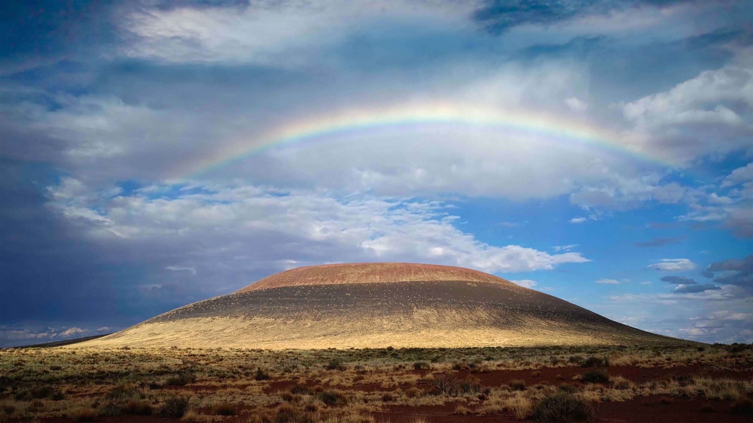 A photograph of blue, cloudy skies with a rainbow over the volanco of Turrell's Roden Crater.