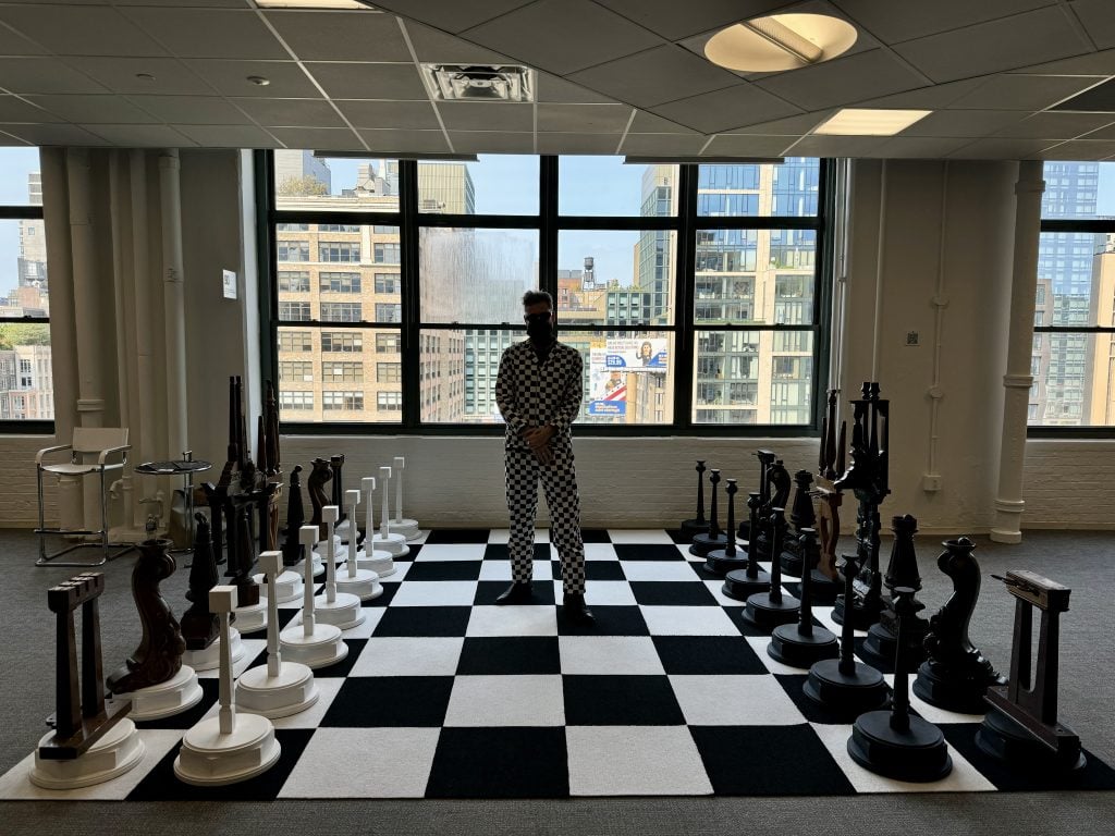 Ryan Bock with his giant chess set made of piano legs, curated by Ki Smith Gallery with Spring Break founders Andrew Gori and Ambre Kelly. He is standing on the giant black and white chess board in a black and white checked suit, standing against a window with views of New York City behind him. 