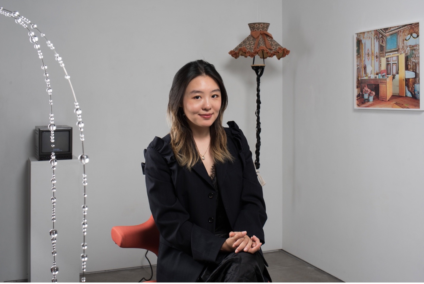 Portrait photograph of gallerist and artist Shihui Zhou sitting in a gallery space dressed in all black, with a floor lamp and a sculpture on a pedestal behind her and a painting on the wall to the right.