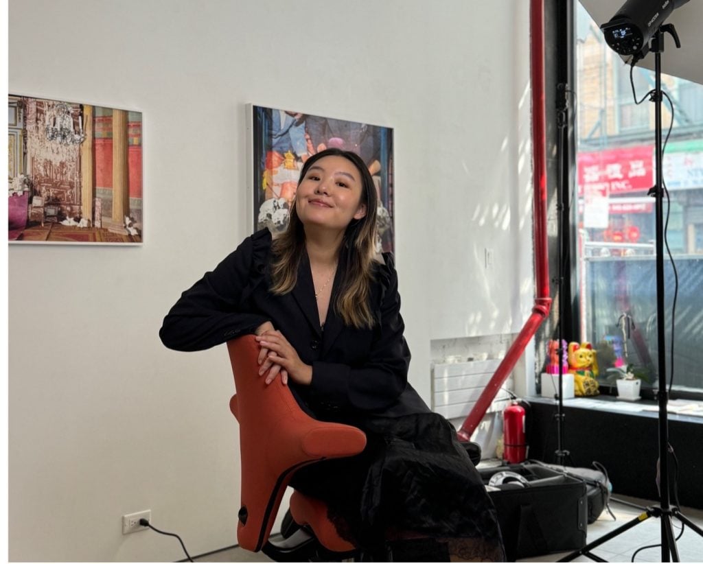 Portrait of artist and gallerist Shihui Zhou sitting in a chair inside the front of the gallery windows with two paintings on the wall behind her.