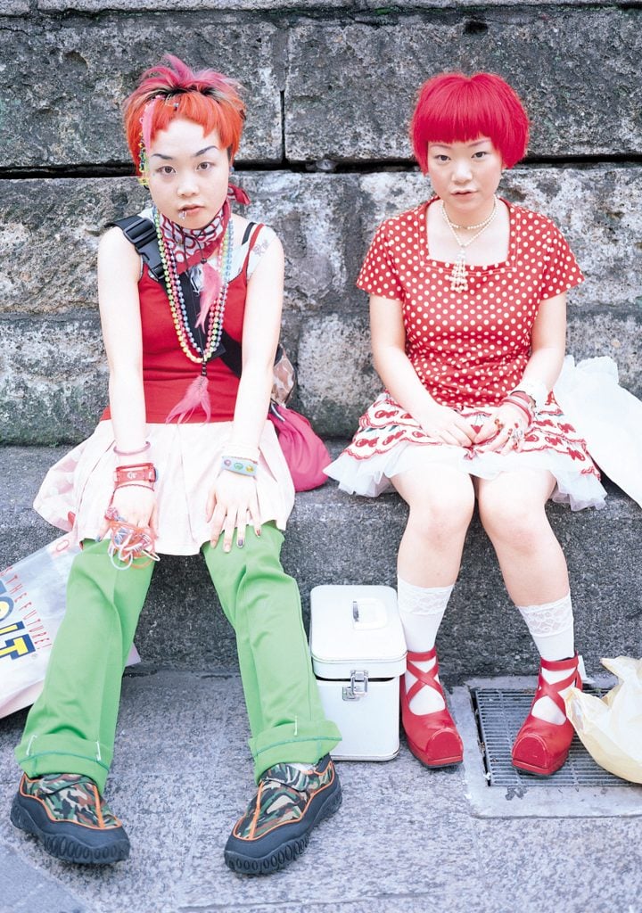 Two individuals wearing bright red pixie cut wigs in cute red and white outfits sitting on a stoop, included in the exhibition we are here