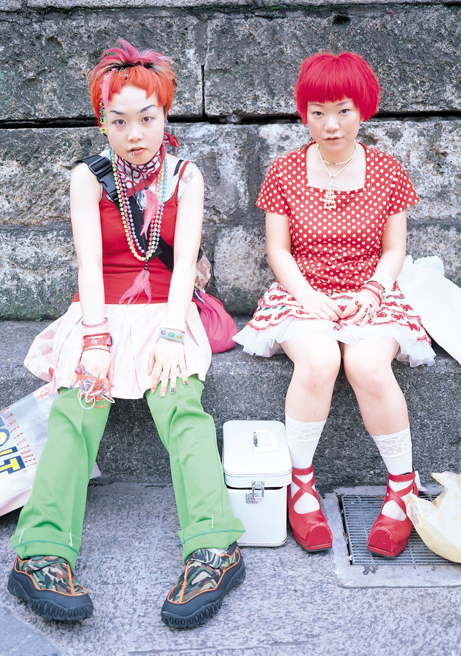 Two individuals wearing bright red pixie cut wigs in cute red and white outfits sitting on a stoop, included in the exhibition we are here