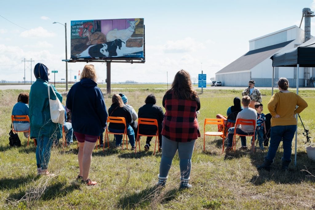 A crowd in a field in front of a billboard by Kandace Creel Falcón titled Guaranteed Income is the G.O.A.T. that shows a painting of goats with the text 