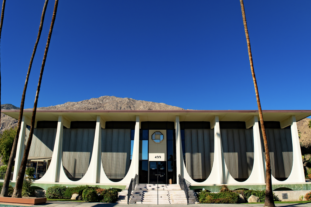 A photograph of a mid-century modern bank beneath blue skies, with palm tree trunks in front of it.