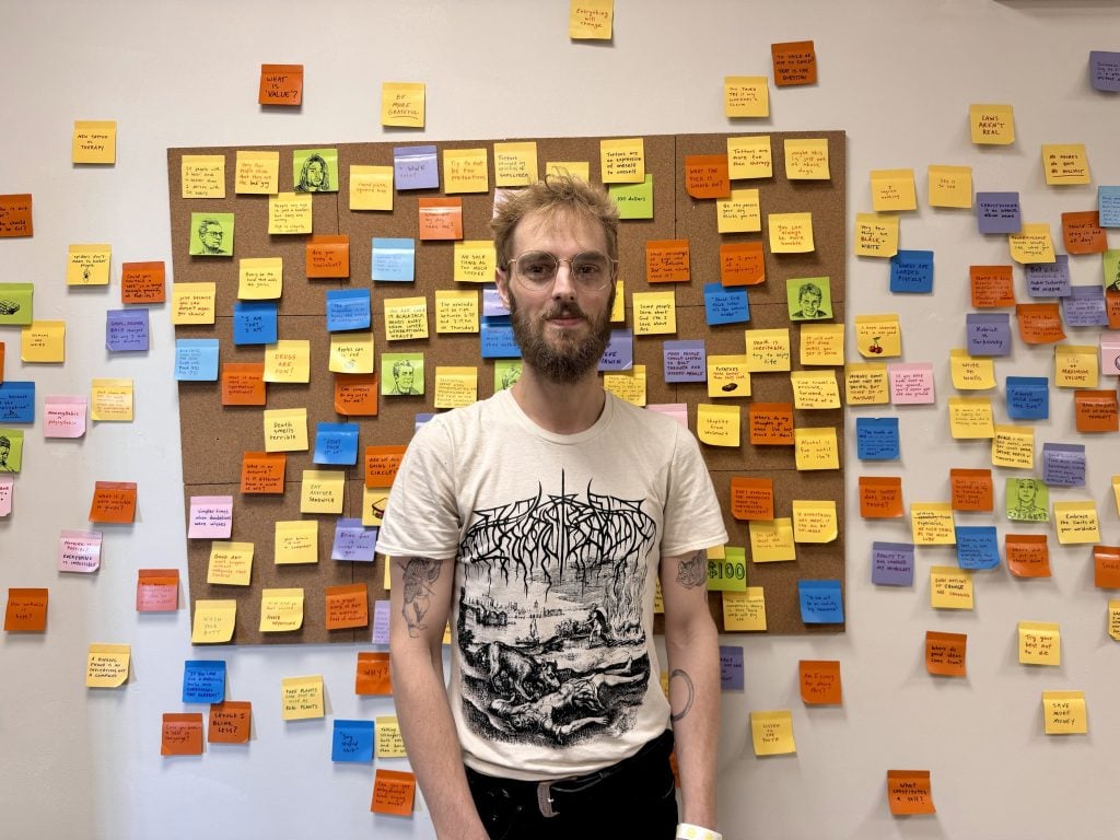 A young white man stands in front of an array of colored Post It notes hanging on a bulletin board and spilling over onto the wall. 