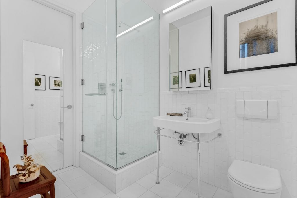 A photograph of a pristine white bathroom with a toilet and shower, from the apartment of John Tancock and Christophe W. Mao.