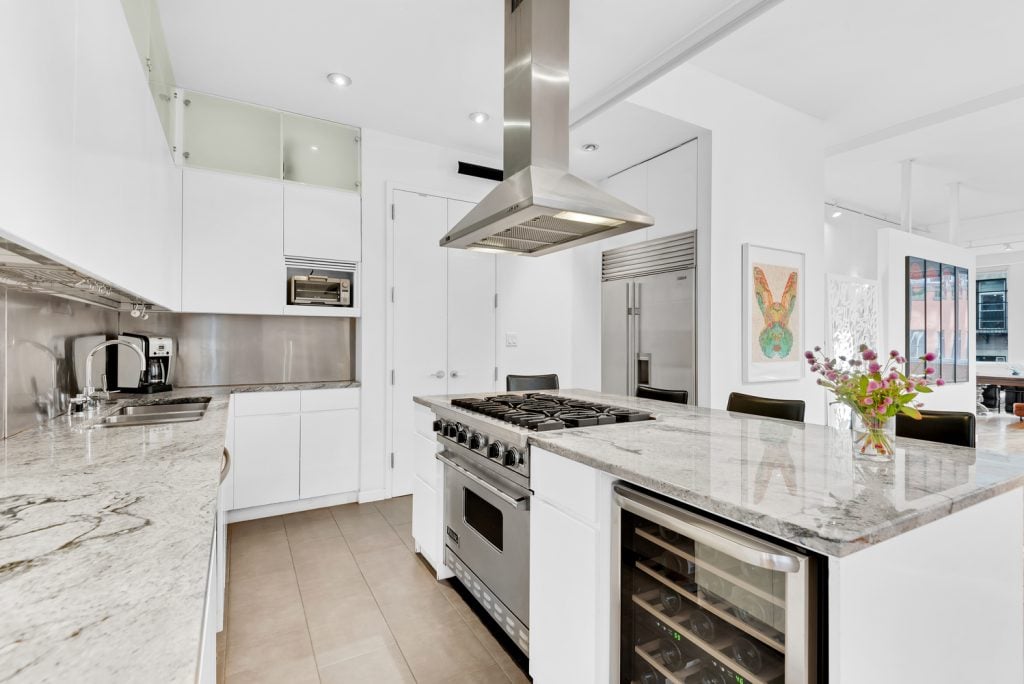 A photograph of a pristine white kitchen with a large stainless steel gas range and a colorful print of a rabbit framed on its white walls in the background.