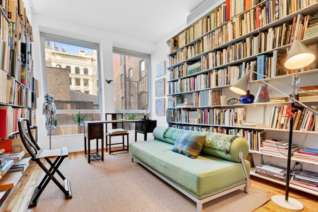 A photograph of a book-lined living room featuring a chrome sculpture and small framed artworks on either side of two large windows, in the apartment of John Tancock and Christophe W. Mao.