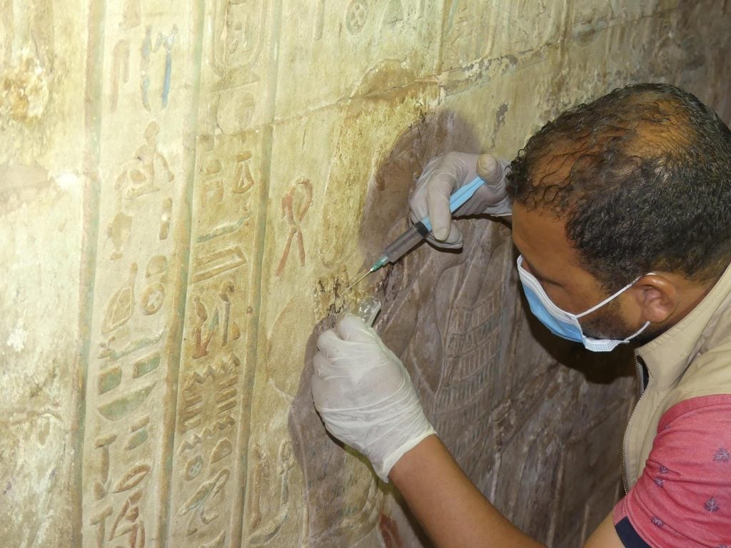 A photograph of a researcher applying pigment to the engraved wall of the ancient Temple of Edfu using a syringe, while wearing gloves and a mask.