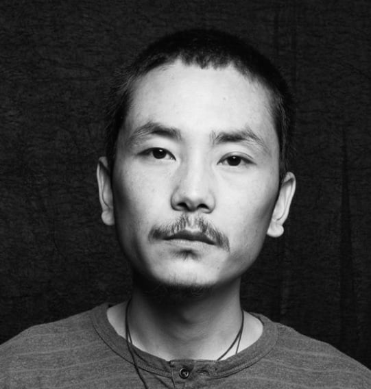 A black and white headshot of the young Tibetan male artist Tenzin Guyrmey Dorjee, depicted on an all black background.